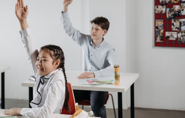 Students actively participating in a classroom setting, seated at desks with raised hands.